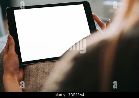 Primo piano di una donna che tiene in mano un tablet con uno schermo bianco vuoto in un ambiente interno accogliente. Mockup perfetto per la progettazione di app, il marketing digitale, il negozio online Foto Stock