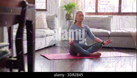 Donna matura seduta a meditare a gambe incrociate nel soggiorno di casa, indossando abiti da yoga su un tappetino rosa Foto Stock