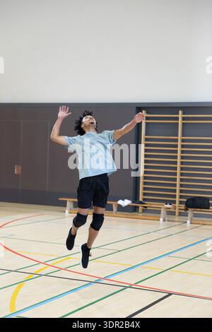 Uomo asiatico, anni 20, che salta su un campo da palestra in legno vicino a sbarre, indossa ginocchiere e scarpe da ginnastica Foto Stock