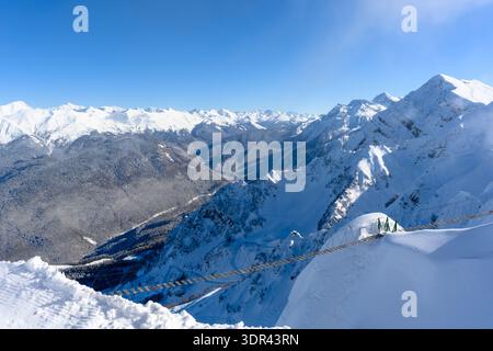 Un ponte sospeso su un precipizio, un'attrazione estrema. Una pittoresca catena montuosa innevata si innalza maestosamente contro un cielo azzurro. Foto Stock