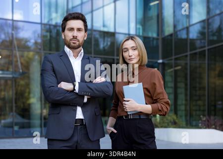 Due partner commerciali sicuri. Un uomo con le braccia incrociate e una donna in possesso di un documento. Si trovano sullo sfondo di un moderno edificio di uffici in vetro. Rappresenta professionalità e lavoro di squadra Foto Stock