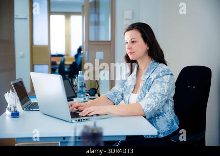 Donna d'affari concentrata che lavora con un notebook alla moderna scrivania. Concetto di produttività professionale, carriera aziendale e stile di vita frenetico. Foto Stock