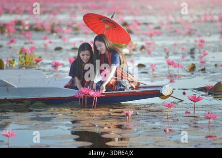 Due giovani donne sedute su una barca con un ombrellone tradizionale che guarda le ninfee rosa, il Mar Rosso del Loto (Talay Bua Daeng), Udon Thani, Thailandia nord-orientale Foto Stock
