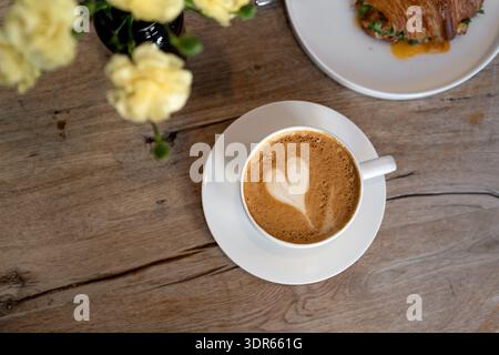 Vista dall'alto di un cappuccino con decorazione a forma di cuore e croissant all'uovo fritto con verdure su un tavolo di legno con fiori in vaso Foto Stock