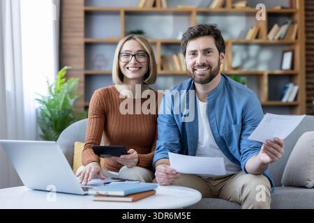 Sorridente giovane coppia seduta su un divano a casa, collaborando alle finanze personali, calcolando le spese e depositando le tasse utilizzando un laptop e un documen Foto Stock