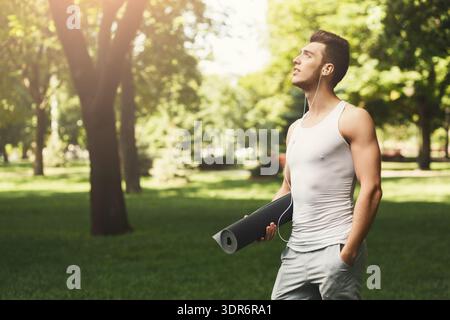 Young Man si trova nel parco con tappetino da yoga e cuffie il giorno della soleggiata Foto Stock