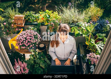 Donna che lavora a distanza con un computer portatile nel lussureggiante giardino con balcone Foto Stock