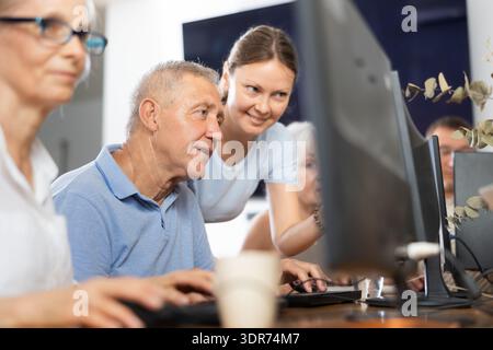 Giovane insegnante di classe informatica per anziani che assiste l'uomo Foto Stock