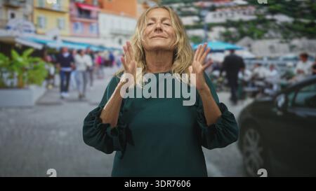 Donna anziana che stringe le mani al petto, gli occhi chiusi con un gesto di preghiera su una strada trafficata con ombrelloni da caffè all'aperto; tranquilla gratitudine. Foto Stock