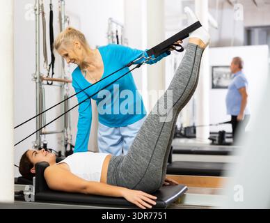 Formatrice femminile esperta che controlla i movimenti di una giovane donna che si esercita sul letto riformatore Pilates in palestra Foto Stock