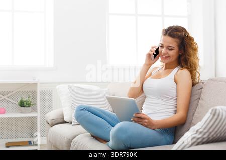 Una donna siede sul divano a parlare al telefono e a utilizzare un tablet in una stanza luminosa durante il giorno Foto Stock