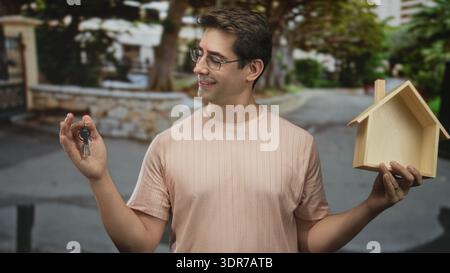 L'uomo tiene le chiavi con la mano sinistra e un modello di casa in legno con la mano destra su una strada, sorridendo e indossando occhiali; orgoglio di proprietà. Foto Stock