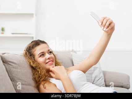 Una giovane donna ama prendere un Selfie mentre si sdraia su un divano a casa nel pomeriggio Foto Stock