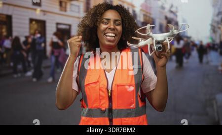 Donna con giubbotto di sicurezza arancione che tiene in mano il drone e fa una pompa a pugno su una strada trafficata della città con pedoni e vetrine sfocate; risultati celebrati Foto Stock