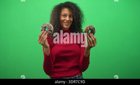 Una ragazza adolescente con capelli ricci tiene due ciambelle cosparse di cioccolato con entrambe le mani contro uno studio di green screen sorridendo; giocosa gioia. Foto Stock
