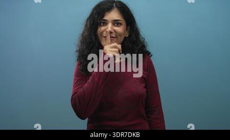 Donna araba che brilla dalle dita alle labbra in studio con sfondo blu con maglione rosso e un leggero sorriso; giocosa segretezza. Foto Stock