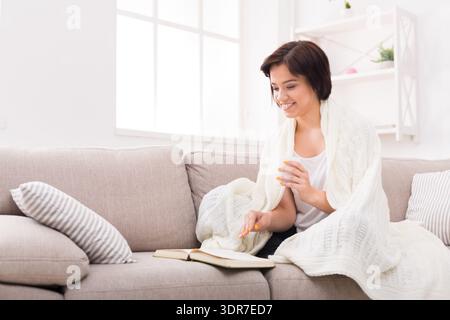 Donna che legge un libro su un divano mentre è avvolta in una coperta e sorride in una stanza luminosa Foto Stock