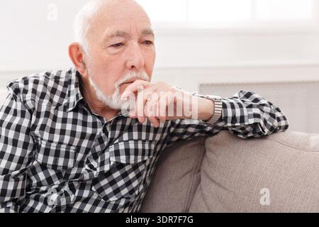 Uomo più anziano in camicia a quadri seduto su un divano con un'espressione premurosa a casa Foto Stock