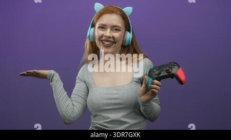 Donna rossa che indossa cuffie azzurre e tiene in mano un gamepad con un gesto palmo in studio; energia giocosa. Foto Stock