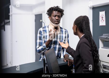 Il giovane uomo nero in braccio al collo controlla con una receptionist per verificare l'appuntamento con il medico. L'amministratore dell'ospedale assiste il paziente maschio ferito, offrendo assistenza e supporto medico. Foto Stock