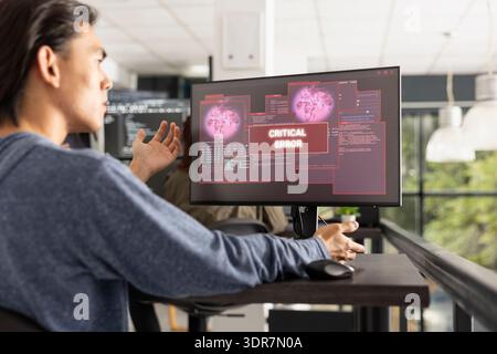 Il programmatore asiatico esamina il monitor del computer mostrando un avviso di errore critico. Il programmatore focalizzato risponde con preoccupazione, sottolineando le problematiche di debug, codifica e risoluzione dei problemi software. Foto Stock