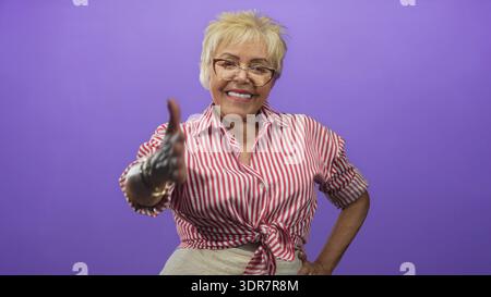 Una donna ispanica anziana sorride e allunga una mano con i guanti per una stretta di mano in uno studio viola; connessione accogliente. Foto Stock