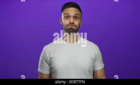 Giovane ispanico con gesti giocosi con il viso gonfiato su un vibrante sfondo viola con una camicia bianca semplice che mostra allegri espressioni Foto Stock