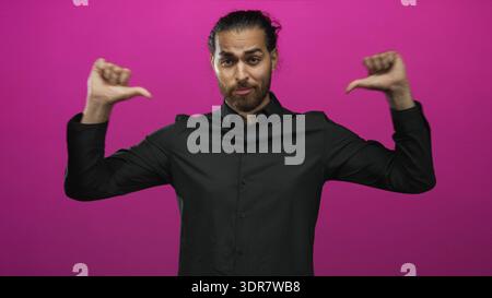Uomo con i pollici che punta al petto, occhi chiusi, barba, camicia nera e panino per capelli in magenta, ambientazione da studio con composizione centrata; orgoglio. Foto Stock