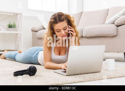 Una giovane donna parla al telefono e usa il laptop mentre si sdraia su un tappeto in una stanza luminosa Foto Stock