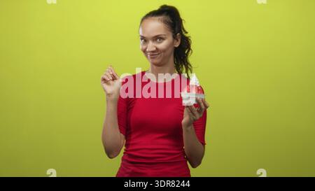 Donna che tiene in mano una tazza di gelato alle fragole con fragole a fette, punta il dito con la mano mentre sorride sullo sfondo verde dello studio; playf Foto Stock
