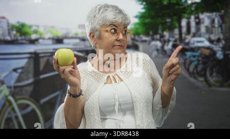 Donna anziana con capelli grigi e occhiali che tiene la mela verde in mano punta il dito su una strada della città sorridente; benessere gioioso. Foto Stock