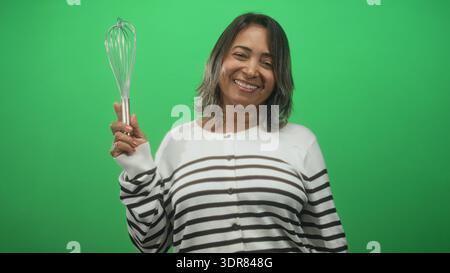 Una donna ispanica sorride e sorride, indossando un maglione a righe, posata davanti a uno schermo verde dello studio per il concetto di cucina; gioia. Foto Stock