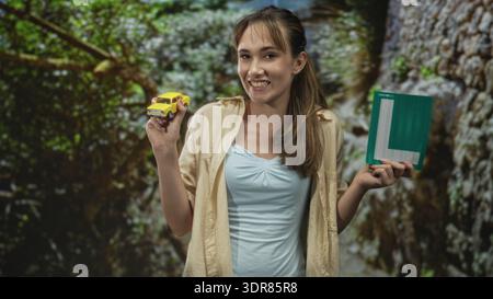 Una giovane donna tiene in mano un'auto giocattolo gialla e un piatto verde per imparare sorridendo e stando in piedi tra alberi e rocce in un parco forestale; imparando sicurezza Foto Stock