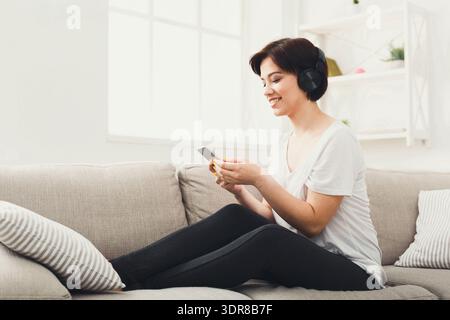 Donna seduta sul divano che ascolta musica sul telefono mentre indossa le cuffie Foto Stock