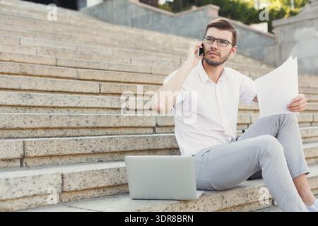 Uomo seduto sui gradini che utilizza il laptop e parla al telefono con i documenti in mano Foto Stock