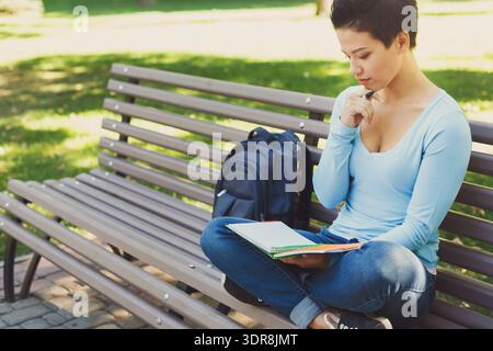 I giovani studiano in un parco mentre sono seduti su una panchina con uno zaino accanto a loro Foto Stock