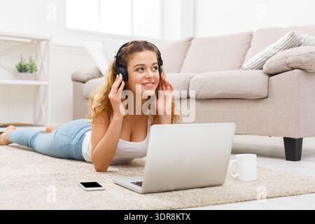 Una giovane donna ascolta la musica a casa mentre è sdraiata sul pavimento con un computer portatile e un telefono nelle vicinanze Foto Stock