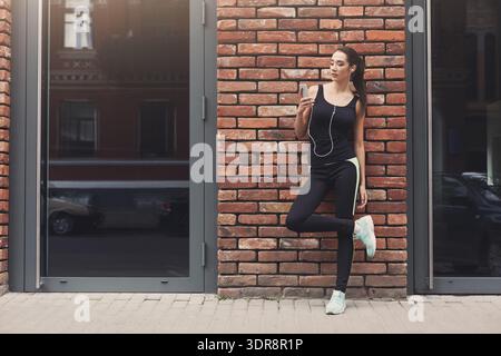 Donna in abiti da ginnastica si appoggia contro il muro di mattoni mentre ascolta musica sullo smartphone Foto Stock