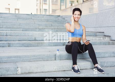 La donna siede sui gradini ascoltando musica mentre si diverte all'aperto e si rilassa in città Foto Stock