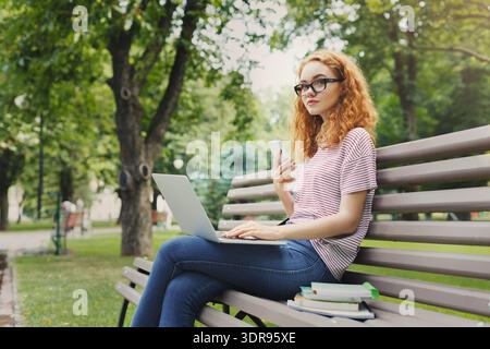 Giovane donna che lavora con un laptop e che controlla il telefono a Park durante il giorno Foto Stock
