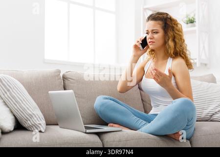 Una donna si siede sul divano utilizzando il telefono e il laptop mentre parla e mostra emozioni Foto Stock