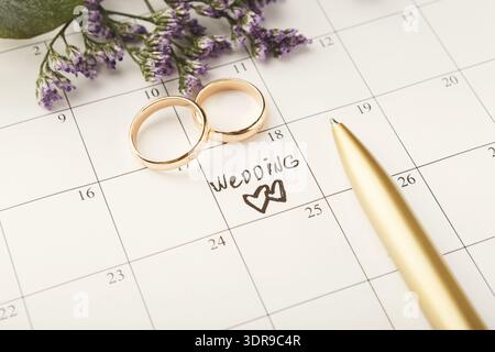 Anelli nuziali sul calendario con una nota per la data di matrimonio e penna nelle vicinanze in un ambiente floreale Foto Stock
