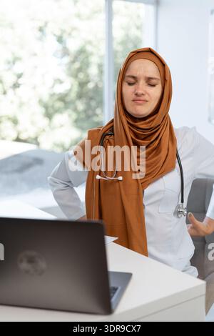 Medico generico arabo professionista. Malsano medico musulmano donna in hijab che soffre mal di schiena inferiore ha lavorato troppo sul portatile in ospedale. Medicina Foto Stock