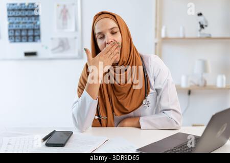 Stanca e sovraccarica di lavoro giovane dottoressa musulmana che indossa un hijab che sbadiglia mentre comunica e lavora nel moderno ufficio della clinica Foto Stock