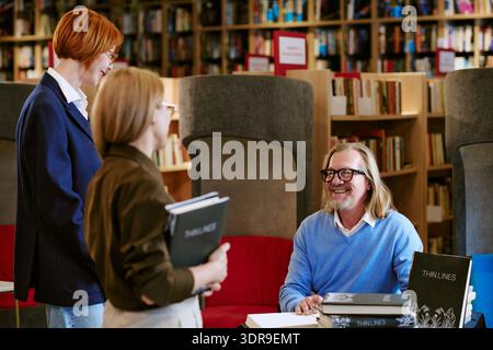 Uomo di mezza età caucasico che firma libri per due donne caucasiche in biblioteca Foto Stock