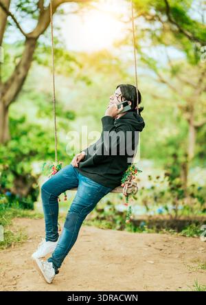 Giovane seduto su un'altalena che chiama al telefono in un giardino. Uomo felice che oscilla al telefono Foto Stock