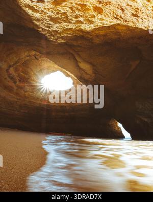 Vista delle travi di sole che si infrangono attraverso la grotta erosa che si apre sulla spiaggia sabbiosa, con dolci onde che si infrangono sulla riva, creando un tranquillo paesaggio costiero Foto Stock