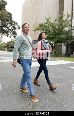 Coppia di mezza età che cammina attraverso la piazza vicino a un edificio in cemento, donna che trasporta una borsa tote rossa e fiori Foto Stock