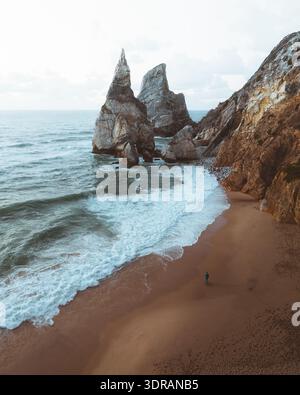Vista aerea delle scogliere spettacolari che incontrano il surf schiumoso, creando un netto contrasto con la spiaggia sabbiosa di Praia da Ursa, Lisbona, Portogallo. Foto Stock