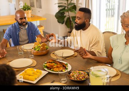 Famiglia multigenerazionale che si tiene per mano e prega prima del pasto, riunita intorno al tavolo da pranzo Foto Stock